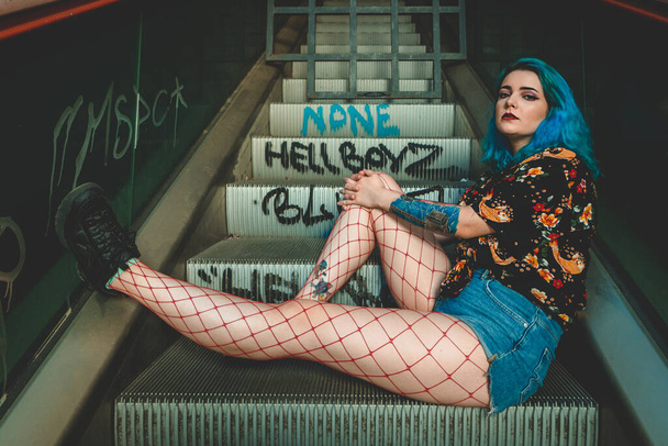 A shallow focus of a stylish blue-haired young woman with fishnet stockings on an idle escalator with graffiti text - Foto, imagen