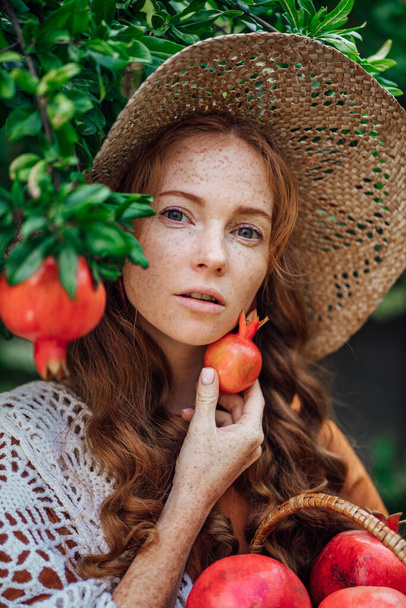 Red pomegranate garden with a happy redhead girl in a hat in an orange dress and white shawl. Close up woman portrait with big red fruits - Photo, Image