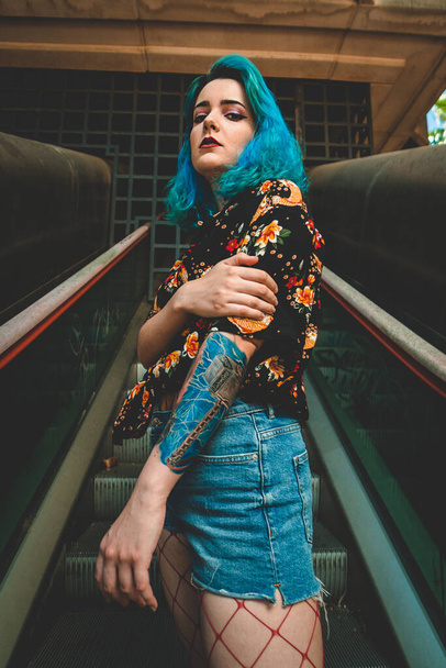 A vertical shot of a stylish blue-haired young woman with fishnet stockings on an idle escalator - 写真・画像