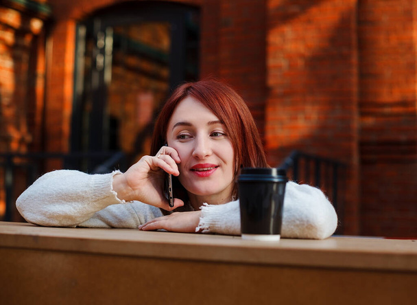 Mulher sorridente apoiou-se na mesa de bar falando no celular. Mulher senta-se no terraço do café enquanto conversa com um amigo em um telefone. - Foto, Imagem