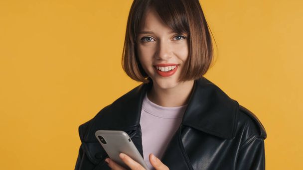 Belle fille brune avec des cheveux de bob en utilisant smartphone regardant joyeusement dans la caméra isolée sur fond jaune - Photo, image