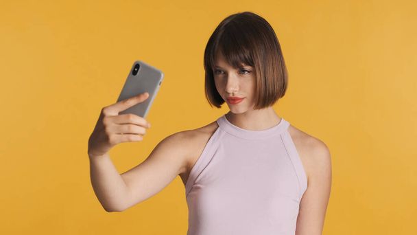 カラフルな背景の上にスマートフォンでセルフィーを作るボブの髪と赤い唇を持つ美しいブルネットの女の子。近代技術の概念 - 写真・画像
