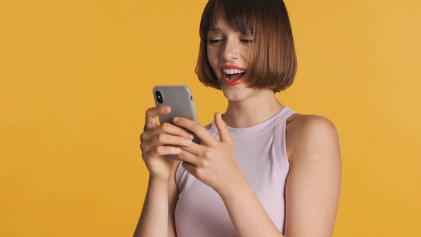 Docela veselá brunetka dívka vychutnat online chatování s přáteli v sociální síti izolované na žlutém pozadí. Šťastný výraz - Fotografie, Obrázek