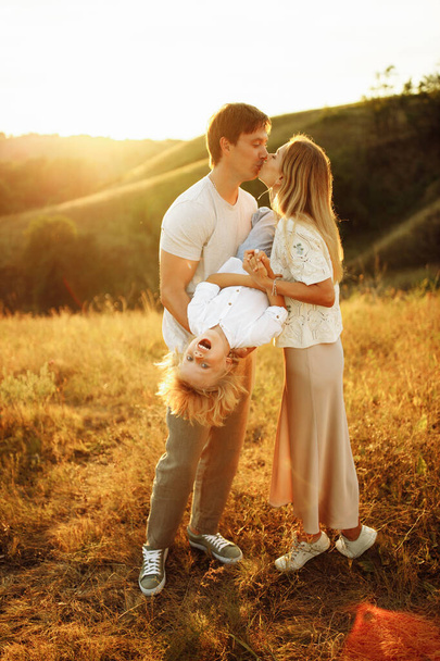 Happy parents with preschool boy at the field, strong father hold joyful son upside down, loving husband gently kiss adorable wife, enjoy spend time together, parenthood concept - 写真・画像