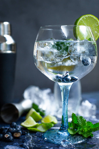 A glass of gin and tonic with mint, lime wedges, blueberries and ice on a grey background - Fotoğraf, Görsel