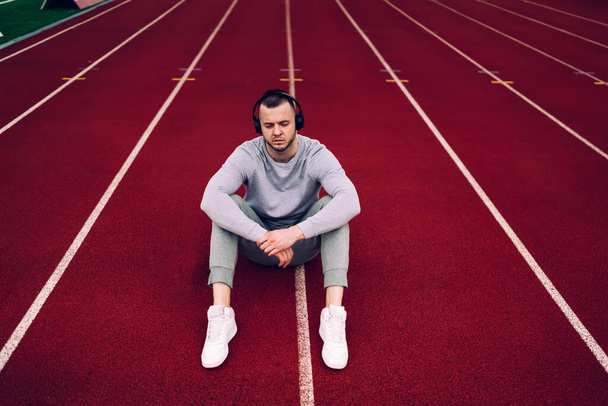 From above of full length sportsman in activewear and sneakers sitting on racetrack and listening to music while resting after workout with closed eyes - Foto, immagini