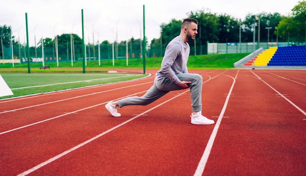 Side view of caucasian male runner stretching lower body muscles for getting better results and training flexible body, handsome young sportsman in activewear having morning workout on stadium - Foto, imagen