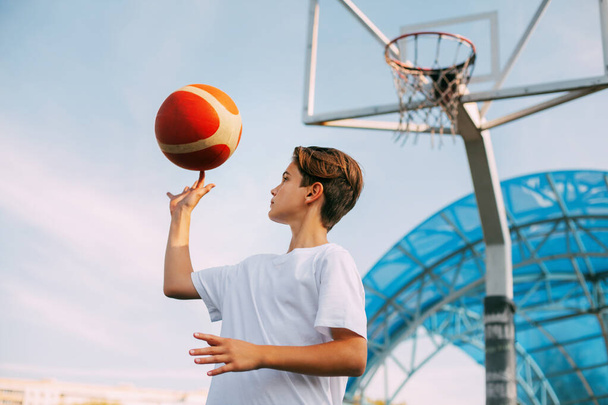 白いTシャツを着た若い10代のバスケットボール選手がバスケットボールコートに立っていて、指でバスケットボールをしている。スポーツと健康的なライフスタイルの概念 - 写真・画像