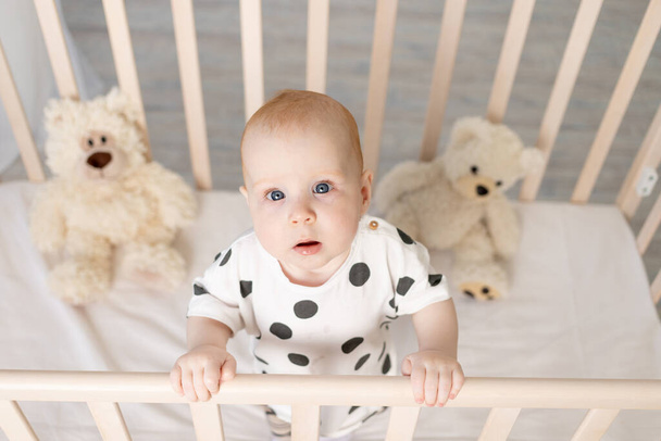 ritratto di un bambino di 8 mesi in piedi in una culla con giocattoli in pigiama in una luminosa stanza dei bambini dopo aver dormito e guardato la macchina fotografica, un luogo per il testo - Foto, immagini