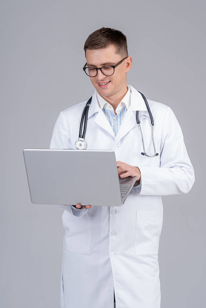 Handsome male medic is standing with laptop in hands. Smiling doctor reading good news on screen. - Photo, Image
