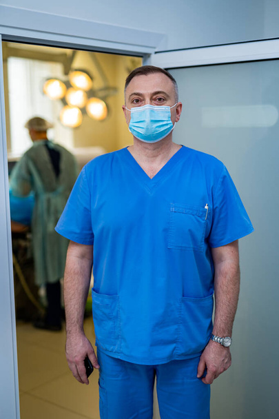 Doctor in mask and scrubs is posing to camera. Medic standing near the surgery room. Closeup. - Foto, immagini