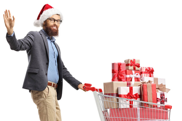 Bearded man with a santa hat waving and walking with a shopping cart full of presents isolated on white background - Photo, Image