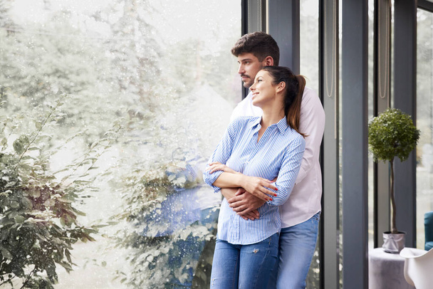 Portrait shot of happy young couple standing by the window and enjoying each others company while snowing outside - Φωτογραφία, εικόνα