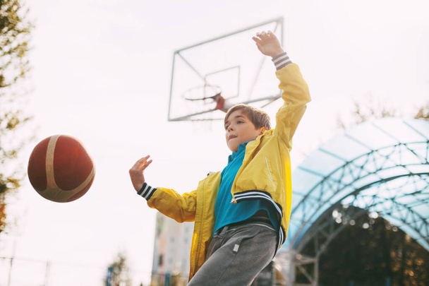 Söpö poika keltainen urheilutakki heittää koripalloa. Lapsi pelaa koripalloa. Urheilu, koulutus, elämäntapa - Valokuva, kuva