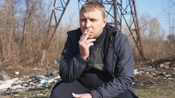 Adult serious man sits squatting and smoking cigarette outdoor. Male person looking into camera and throwing out a cigarette butt. Concept of unhealthy habit and addiction. Close up Slow motion. - Photo, image