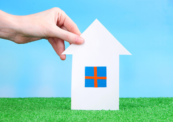 Little paper house in hand close-up, on green grass, on blue sky background - Photo, Image