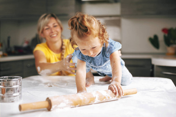 A cheerful little girl, soiled in flour, crawls away from her mother on the table. Blurred background, space for text - Foto, Imagen