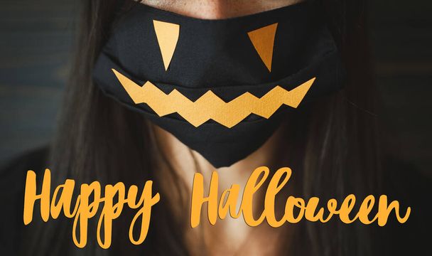 Happy Halloween κείμενο για νεαρή γυναίκα σε μαύρο πρόσωπο μάσκα με jack o lantern χαμόγελο σε σκούρο ξύλινο φόντο. Απόκριες 2020, να είστε ασφαλείς κατά τη διάρκεια του ιού της στέψης. Χειρόγραφο σήμα, εποχιακή ευχετήρια κάρτα - Φωτογραφία, εικόνα