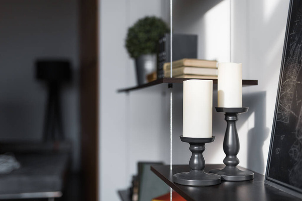 two candles in black wooden candlesticks stand on a shelf indoors on a Sunny day in a dark room design - Photo, image