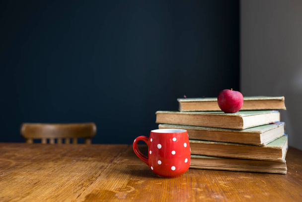 A stack of large, old, rag-bound books lie on a wooden table against a dark blue wall. Pen and notebook. Concept - back to school. Online training. Copy space. Select focus. Red cup with white polka dots with tea. - 写真・画像