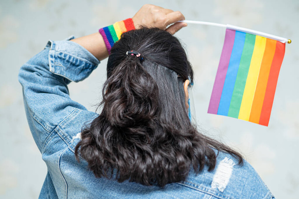 Azjatka w masce i tarczy na twarz dla ochrony wirusa covid-19 z tęczową flagą, symbolem miesiąca dumy LGBT świętować coroczne w czerwcu towarzyskie gejów, lesbijek, biseksualistów, transseksualistów, praw człowieka. - Zdjęcie, obraz