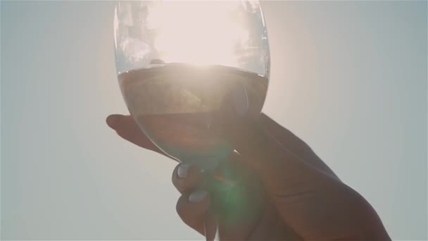 Vrouwen hand zwaaien goud witte wijn in glas op gedeconcentreerde achtergrond met stralen van zonlicht - Video