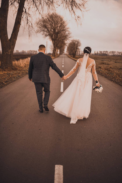 新婚夫婦はアスファルトの道路に沿って手を握っている - 写真・画像