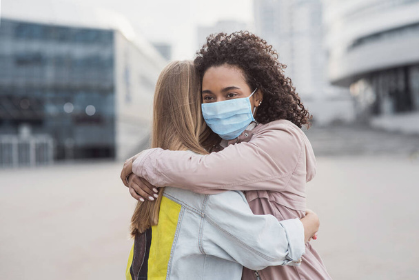 Meeting of two happy friends hugging in the street wearing protective face mask. Multiethnic young women embrace each others in a city during corona virus pandemy - Фото, зображення