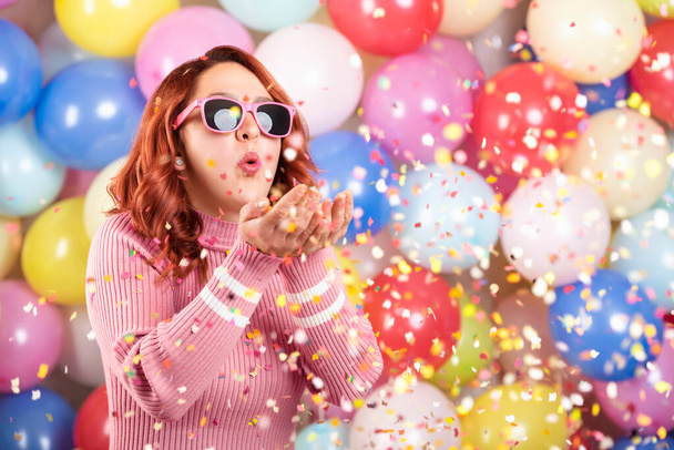 Redhair young woman parties alone, blowing confetti on multicolored balloons background. Celebrating alone at home concept. Happy girl alone at party. - Photo, Image
