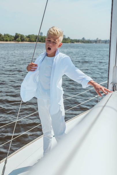 Serious teen handsome blonde boy in white t-shirt and shirt singing on the yacht. Education, lessons, studying, skills, growing up concept - Foto, immagini