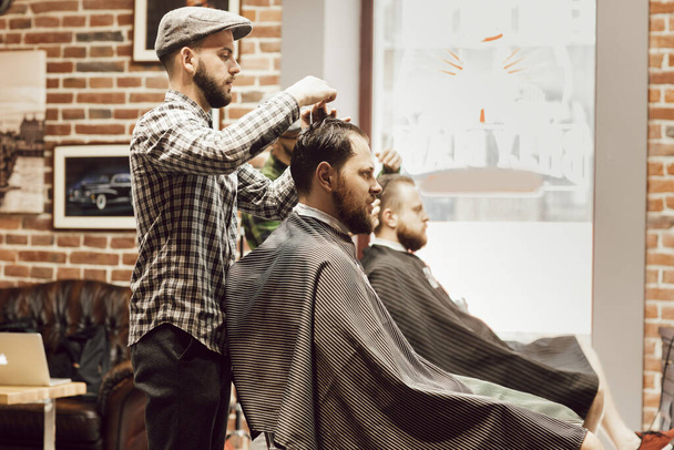 Berber dükkanında saç kesimi. Genç sakallı adam berber dükkanında sandalyede otururken saçını kestiriyor. Kuaför saçı makas ve tarakla keser. - Fotoğraf, Görsel