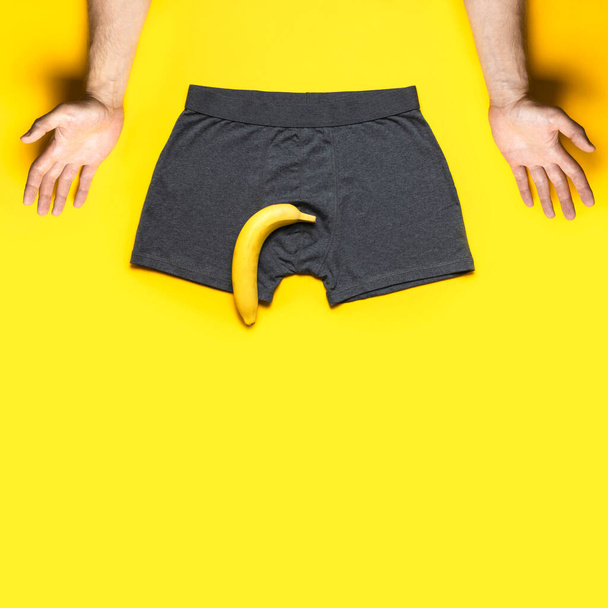 男性の能力の問題の創造的な概念。黄色の背景フラットレイ上のグレーのパンティー新鮮なバナナ男性の手の上ビュー。男性の性的機能障害,健康上の問題.勃起不全の影響. - 写真・画像