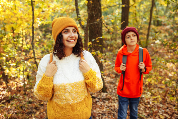 Una giovane donna e un ragazzo passeggiano nel parco in autunno. Donna che indossa cappello arancione e maglione e escursione adolescente nella foresta con zaini durante la stagione autunnale. Concetto di esplorazione della natura - Foto, immagini