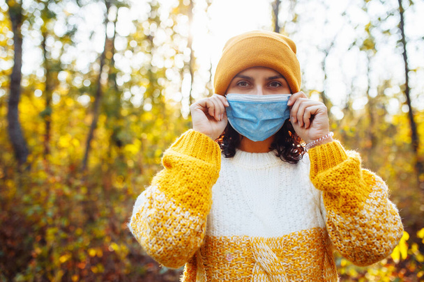 Mujer joven con un sombrero naranja y suéter se quita la máscara médica estéril en el bosque de otoño. La hembra respira aire fresco en el parque de otoño durante la pandemia del coronavirus covid-19. Concepto sanitario - Foto, Imagen