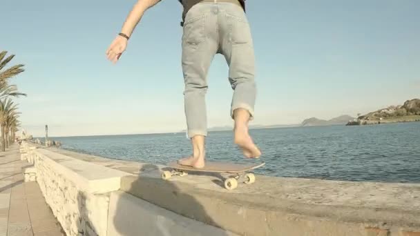 Niño vistiendo jeans azules patinando descalzo en el muelle con el mar al fondo - Imágenes, Vídeo