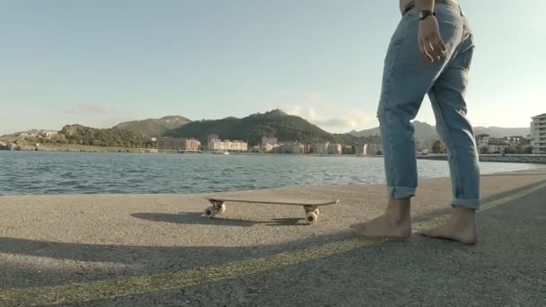 Joven sin camisa y vaqueros azules agarra su monopatín y se sienta frente al mar en la soleada tarde de verano - Imágenes, Vídeo