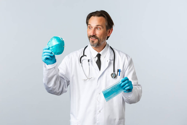 Covid-19, prevence viru, zdravotnických pracovníků a koncepce očkování. Nadšený lékař středního věku v bílém plášti a rukavicích, ukazuje dva PPE, dívá se na potěšený respirátor, drží masku - Fotografie, Obrázek