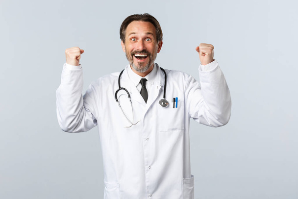 Covid-19, sepelvaltimotautiepidemia, terveydenhuollon työntekijät ja pandemia. Helpotettu onnellinen mieslääkäri saa hyviä uutisia, nostamalla kädet ylös juhlassa ja hymyilemällä tyytyväinen, voittoisa - Valokuva, kuva
