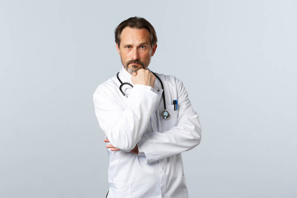 Covid-19, coronavirus outbreak, healthcare workers and pandemic concept. Serious-looking professional male doctor in white coat, touch beard and looking thoughtful camera, treating patients - Photo, Image