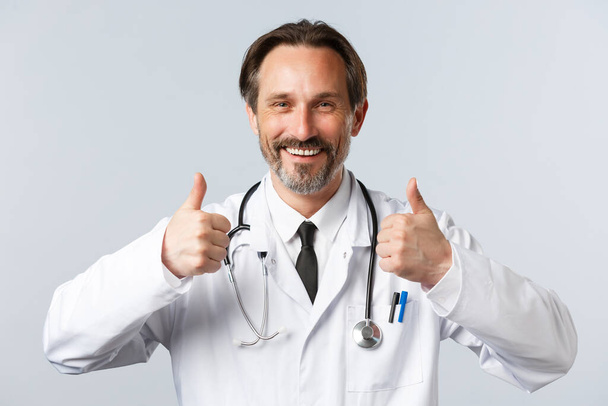 Covid-19, prevence viru, zdravotnických pracovníků a koncepce očkování. Hezký spokojený lékař v bílém plášti, s úsměvem a palcem nahoru ve schválení, doporučujeme a garantujeme kvalitu - Fotografie, Obrázek