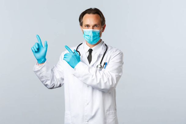 Covid-19, preventing virus, healthcare workers and vaccination concept. Confident serious male doctor in white coat, medical mask and gloves pointing top left at banner with information - Photo, Image