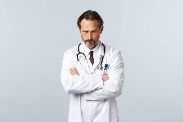 Covid-19, ξέσπασμα του ιού της στέψης, επαγγελματίες υγείας και πανδημία. Σοβαρή απογοητευμένη αρσενικό γιατρό σε λευκό παλτό, σταυρό στο στήθος μπράτσα και να δούμε κάτω από το μέτωπο judgemental - Φωτογραφία, εικόνα