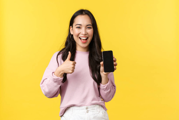 Az emberek érzelmei, életmód szabadidő és szépség koncepció. Mosolygó ázsiai nő 20-as, mutatja okostelefon kijelző, ajánlott alkalmazás vagy mobil játék, mutatja hüvelykujj-up jóváhagyással, sárga háttér - Fotó, kép