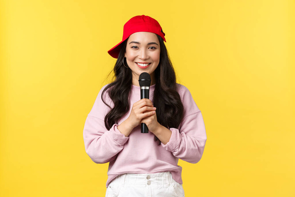 Az emberek érzelmei, életmód szabadidő és szépség koncepció. Aranyos mosolygós ázsiai nő előadóművész ének dal piros sapkában, gazdaság mikrofon, mint a beszéd vagy éneklés rap, álló sárga háttér - Fotó, kép