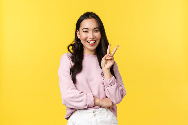 People emotions, lifestyle and fashion concept. Stylish korean girl in casual outfit showing peace sign and smiling silly, having good summer vacation, enjoying leisure, stand yellow background - Foto, Bild