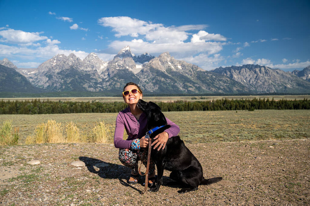 A nő öleli és tartja a fekete labrador retriever kutya előtt a Grand Teton Nemzeti Park hegyek Jackson Wyoming ahogy a kutya nyalogatja az arcát - Fotó, kép