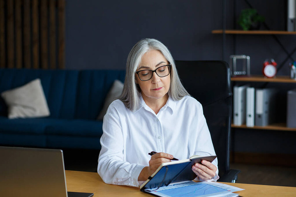 Portrait of beautiful smiling elderly woman studying online. Online education, remote working, home education. Grey-haired senior woman writing in notebook. Mature people study courses online - Foto, imagen