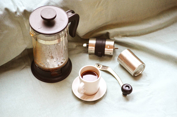 Dampfende nasse französische Presse, Kaffeetasse mit dunklem heißen Espresso auf Untertasse und kleine Kaffeemühle aus Metall auf hellgrünem Stoff. Stillleben. Frischen Kaffee zu Hause zubereiten. - Foto, Bild
