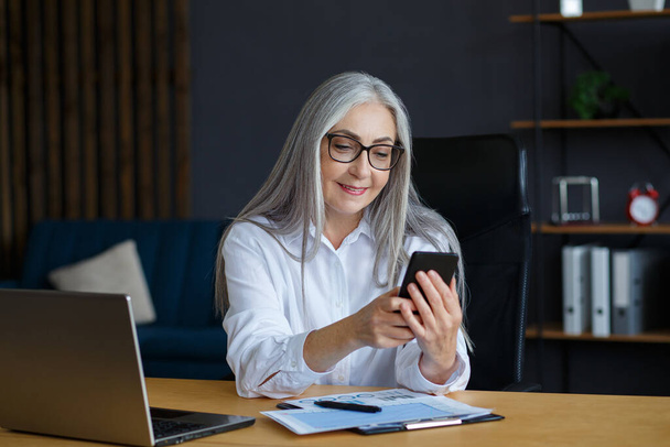 Senior grey-haired beautiful woman using smartphone and smiling. Happy businesswoman using mobile phone apps, texting message, browsing internet, shopping online. Mature people with mobile devices - Φωτογραφία, εικόνα
