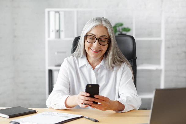 Senior grey-haired beautiful woman using smartphone and smiling. Happy businesswoman using mobile phone apps, texting message, browsing internet, shopping online. Mature people with mobile devices - Φωτογραφία, εικόνα